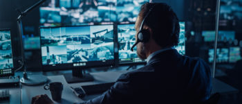 Male Officer Works on a Computer with Surveillance CCTV Video in a Harbour Monitoring Center with Multiple Cameras on a Big Digital Screen. Employees Sit in Front of Displays with Big Data.